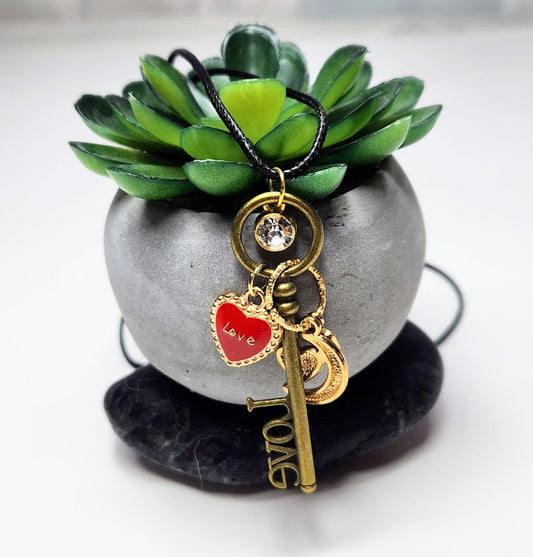 Handcrafted adjustable corded necklace with a vintage-style key, a sparkling gem, and a red heart charm displayed against a artificial green plant backdrop.