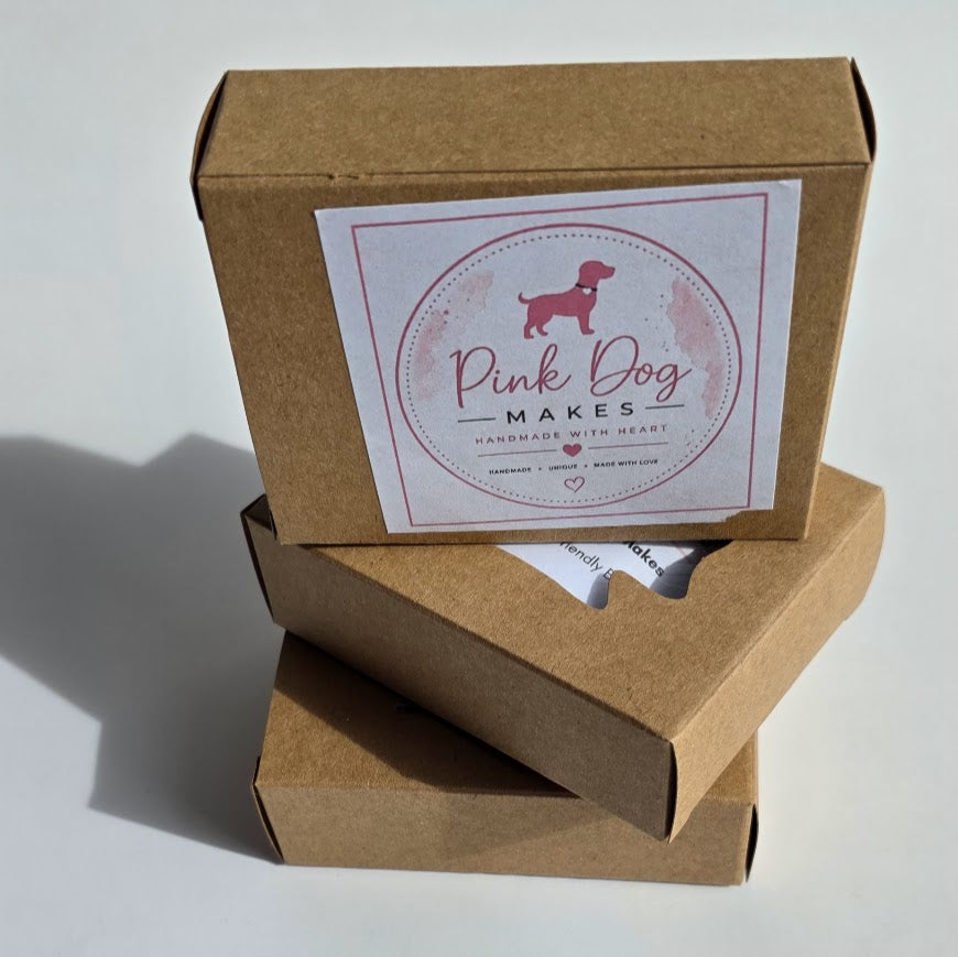 Three stacked brown kraft paper gift boxes on a white background, with the top box featuring a white label that reads 'Pink Dog Makes' with a pink dachshund silhouette.