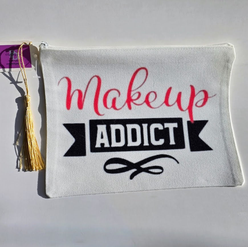 A white canvas makeup bag with a gold tassel and a purple hang tag, featuring the text 'Makeup ADDICT' in red and black on the front, set against a plain light background.