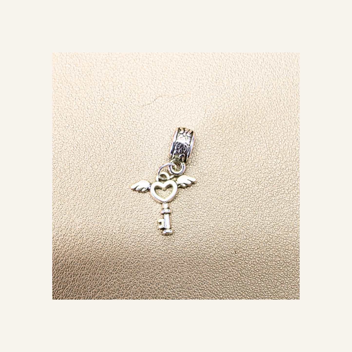 A silver key charm with wing and heart details, displayed on a textured beige surface.
