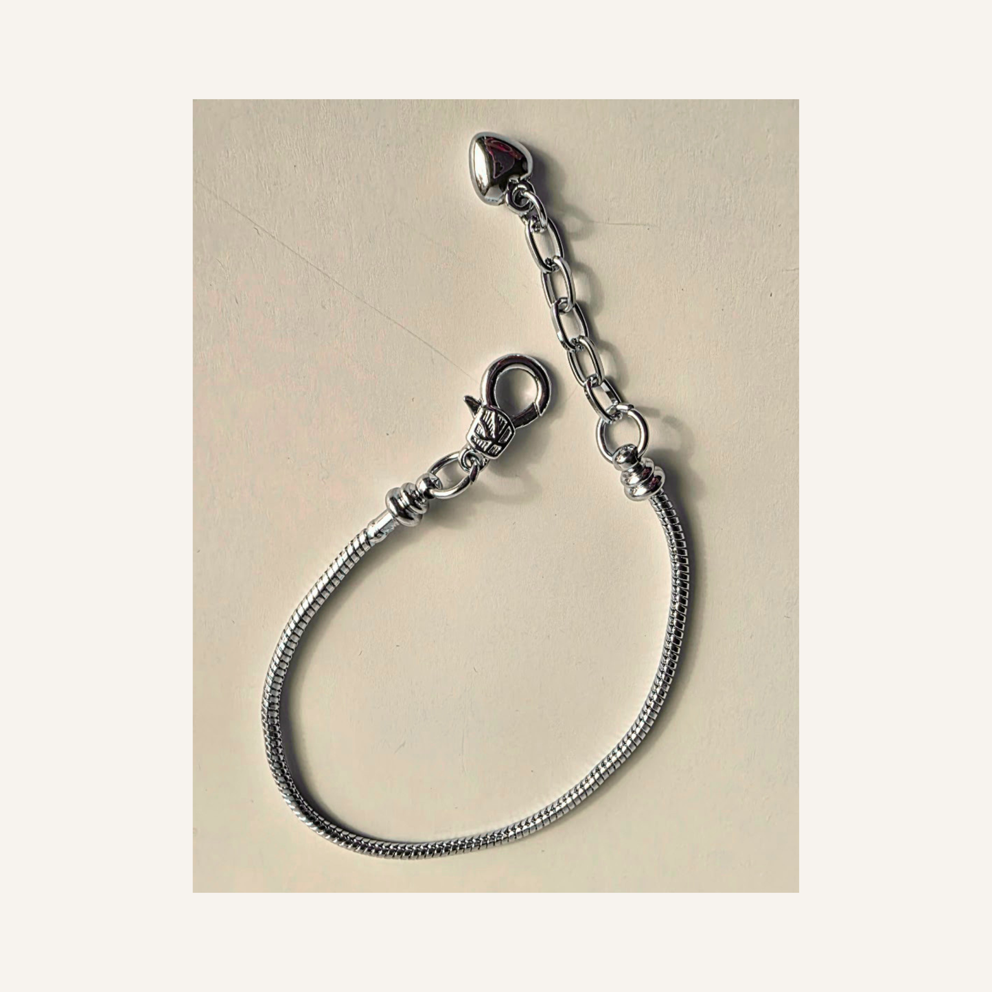 A silver snake chain bracelet with a removeable lobster clasp and an adjustable extension chain featuring a small heart-shaped charm, displayed on a light beige background.