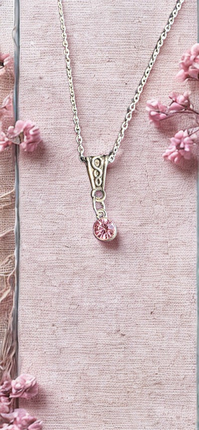 A silver-toned necklace with a decorative bail and a small round pink gemstone pendant, displayed on a textured pink fabric background with scattered pink flowers.