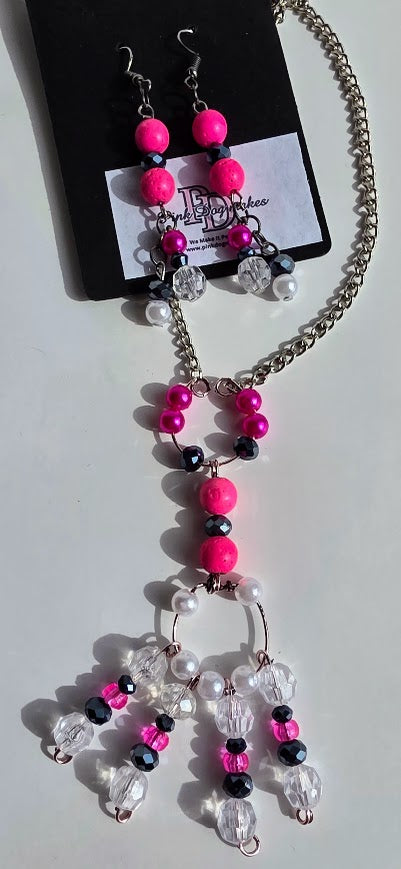 A beaded necklace and matching drop earrings displayed on a black card. The jewelry features bright hot pink beads, black beads, clear faceted beads, and white pearl-like beads on a silver-tone chain.