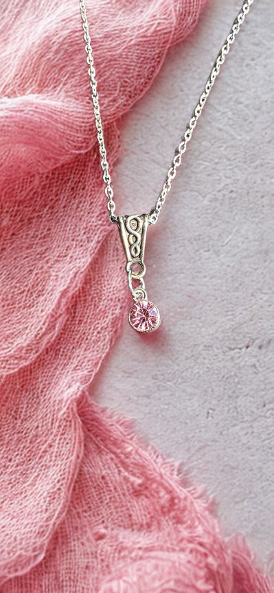 A silver-toned delicate chain necklace with an ornate pendant featuring a small dangling pink gemstone, styled on a textured light gray surface with a soft pink fabric drape.