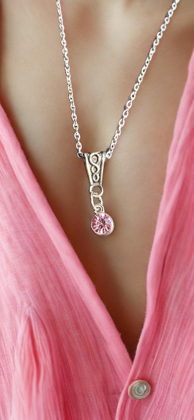 A close-up of a silver pendant necklace with a pink faceted gemstone, worn by a model wearing a pink button-down shirt.