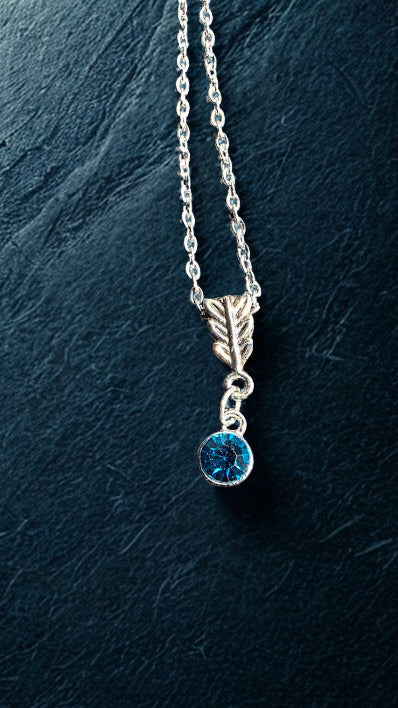 A silver necklace featuring a leaf-shaped charm and a dangling round blue gemstone, displayed against a dark textured slate background.
