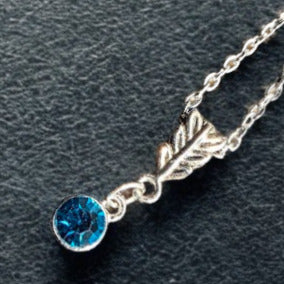 A silver necklace featuring a round, faceted blue gemstone pendant and a small leaf-shaped charm, displayed on a dark textured background.