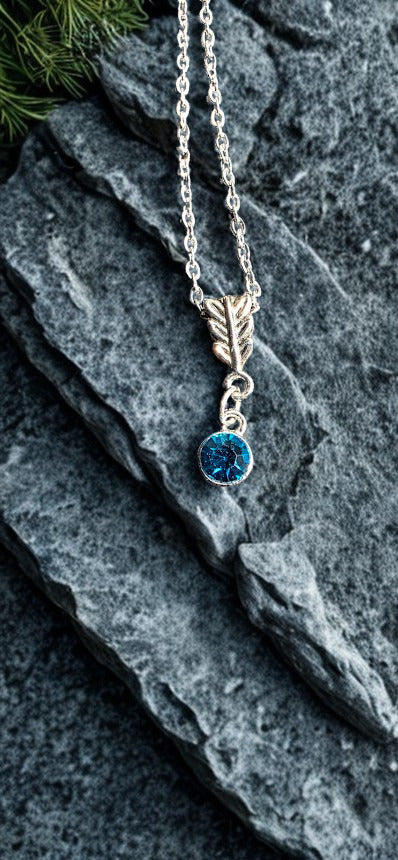 A silver-toned necklace with a leaf-shaped charm and a round blue gemstone pendant, displayed on dark textured slate with a small sprig of greenery at the top left.