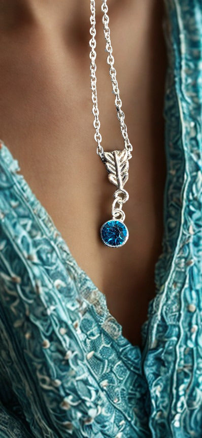 A close-up of a silver necklace with a leaf-shaped pendant and a round blue gemstone, worn on a model's neck over a textured teal top.