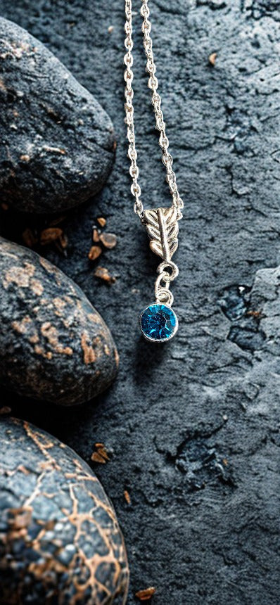 A silver-toned necklace with a leaf-shaped pendant and a round blue gemstone, displayed on dark textured rocks.