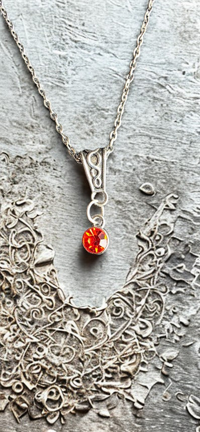 A silver pendant necklace with a small, round, vibrant orange-red gemstone, displayed on a textured gray stone surface next to a silver filigree collar.