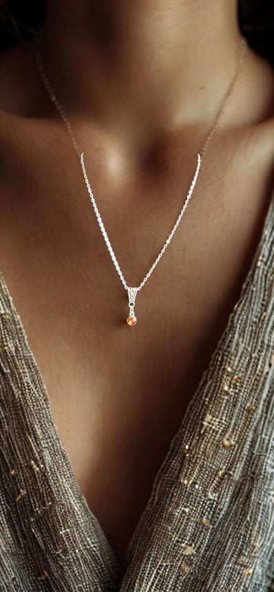 A close-up of a model's neck and chest wearing a delicate silver chain necklace with a small orange teardrop pendant, paired with a shimmering metallic bronze V-neck top.