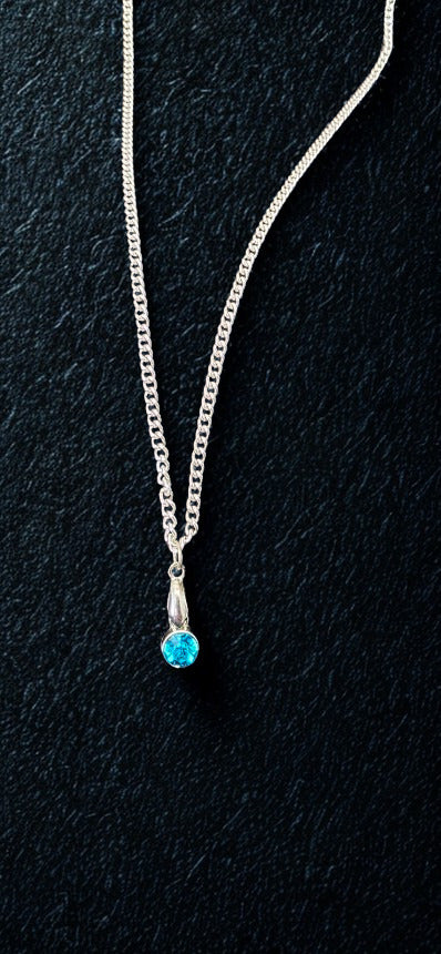 A silver chain necklace with a small silver pendant holding a round-cut bright blue gemstone, displayed on a dark textured background.