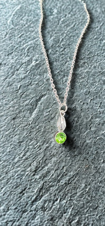 A delicate silver chain necklace with a small silver pendant featuring a vibrant green gemstone, displayed on a textured gray slate surface.