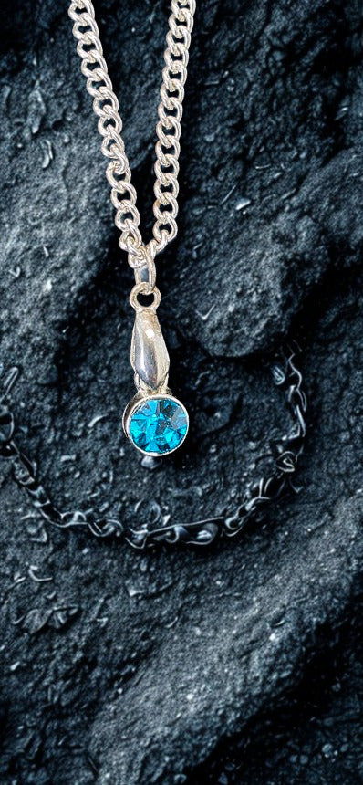 A silver chain necklace with a teardrop-shaped silver pendant holding a round, vibrant blue gemstone, displayed on a textured black rock surface.