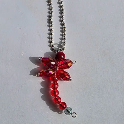 A red dragonfly pendant necklace made of faceted red beads and a silver ball chain, photographed on a plain light background.
