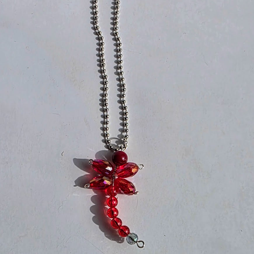 A red dragonfly pendant necklace made of faceted red crystals and red beads on a silver ball chain, photographed on a light gray surface.