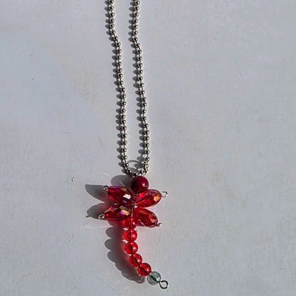 A red dragonfly pendant necklace made of faceted red crystals and red beads on a silver ball chain, photographed on a light gray surface.
