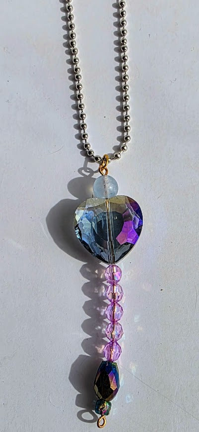 A beaded necklace featuring a large heart-shaped iridescent blue glass bead, a clear faceted bead, a strand of pink beads, and an iridescent dark bead, suspended from a silver ball chain.