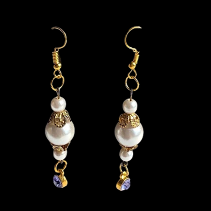 A pair of gold and white drop earrings with white pearl-like beads, gold filigree details, and small purple crystal charms, shown against a solid black background.