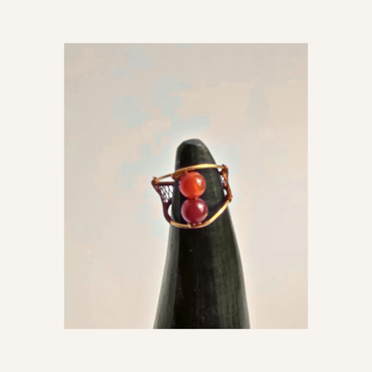 A gold ring with two red agate gemstone beads, displayed on a black cone stand against a light beige background.