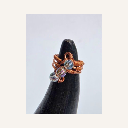 A handmade copper wire ring with three clear faceted beads, displayed on a black cone stand against a light gray background.