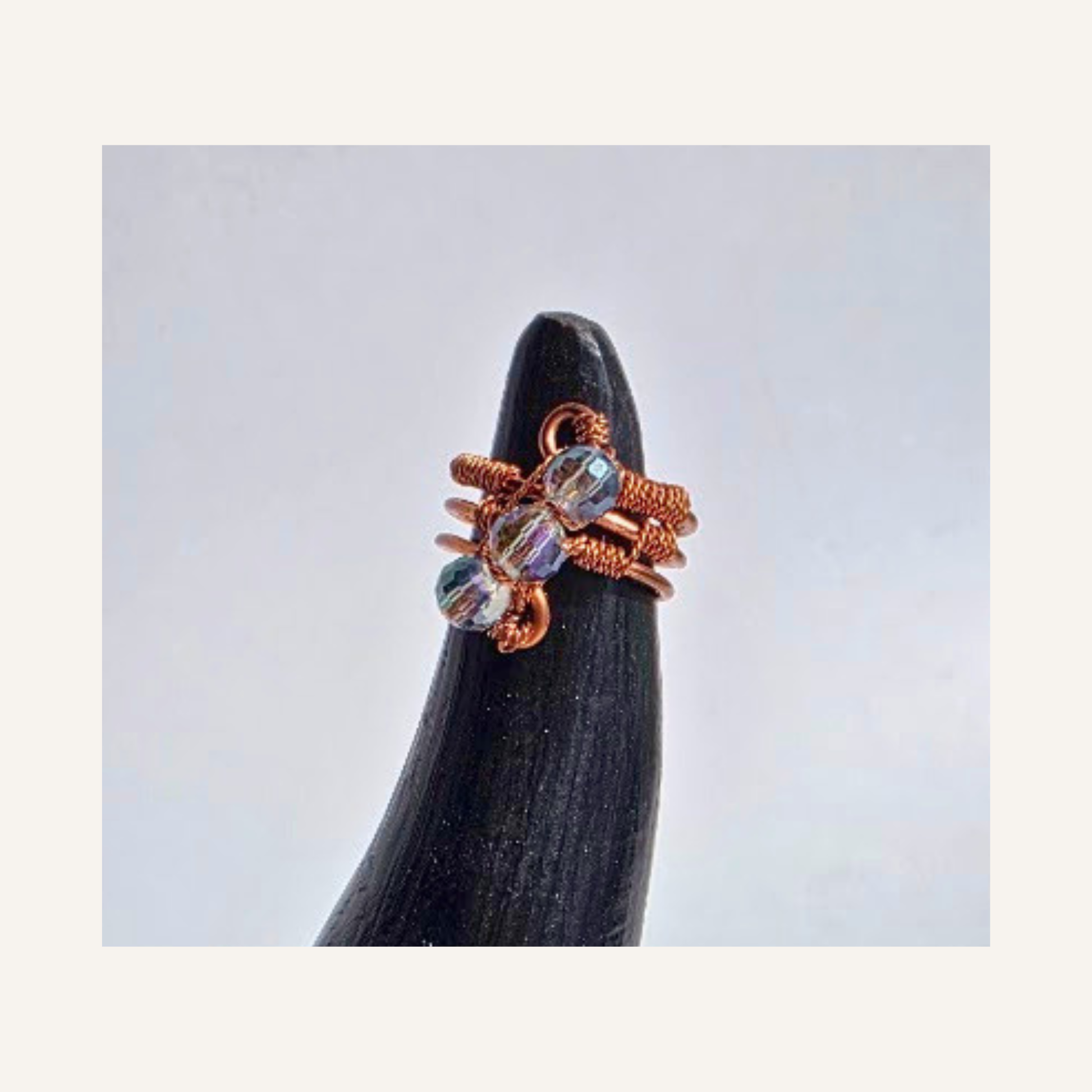 A copper wire wrapped ring with iridescent glass beads, displayed on a black cone stand against a light gray background.
