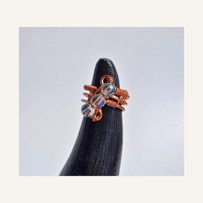 A copper wire wrapped ring with iridescent glass beads, displayed on a black cone stand against a light gray background.