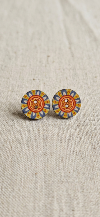 A pair of round stud earrings with a colorful mandala pattern in orange, yellow, blue, and white, resting on a textured beige linen background.