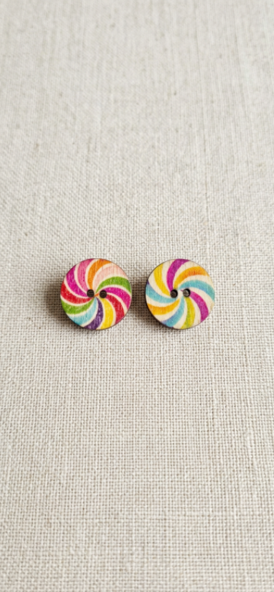 A pair of round stud earrings with a colorful spiral candy-lollipop pattern, displayed on a textured beige linen background.