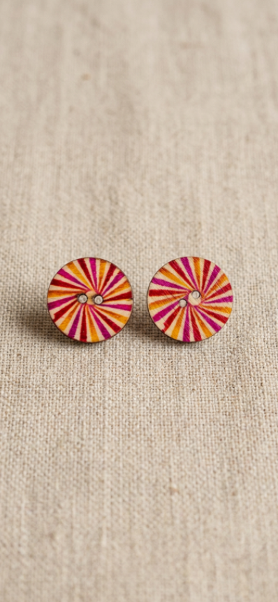 A pair of round stud earrings with a colorful radial sunburst pattern in pink, orange, and yellow, displayed on a textured beige linen background.