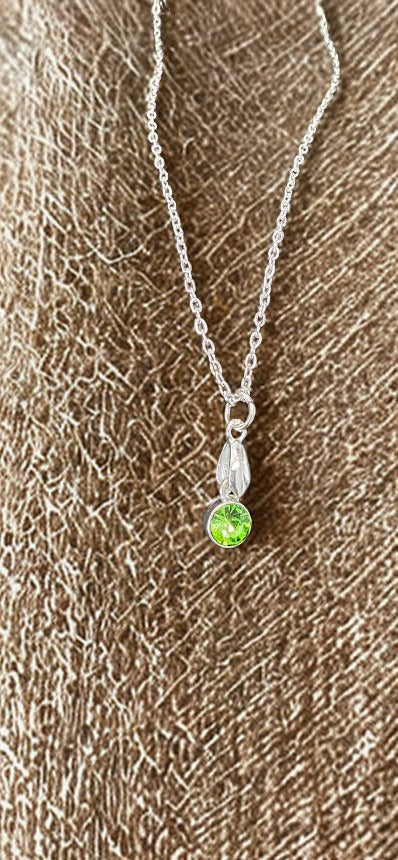 A silver chain necklace with a small silver pendant featuring a round, bright green gemstone, displayed on a textured brown fabric background.