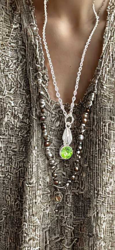 A close-up of a silver-toned necklace with a leaf-shaped pendant featuring a bright green stone, layered over dark brown and gray beaded strands on a textured gray top.