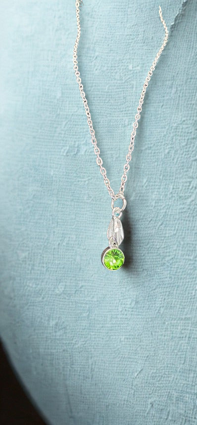 A silver chain necklace with a small silver pendant featuring a round green gemstone, displayed on a textured light blue surface.