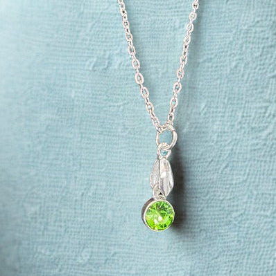 A silver chain necklace with a small silver pendant featuring a round green gemstone, displayed on a textured light blue surface.