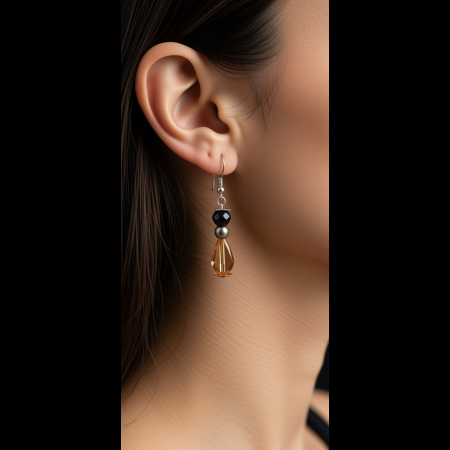 A close-up of a person's ear wearing a dangling earring with a silver-toned metal hook, a small black bead, a silver spacer bead, and a faceted amber-brown teardrop charm.