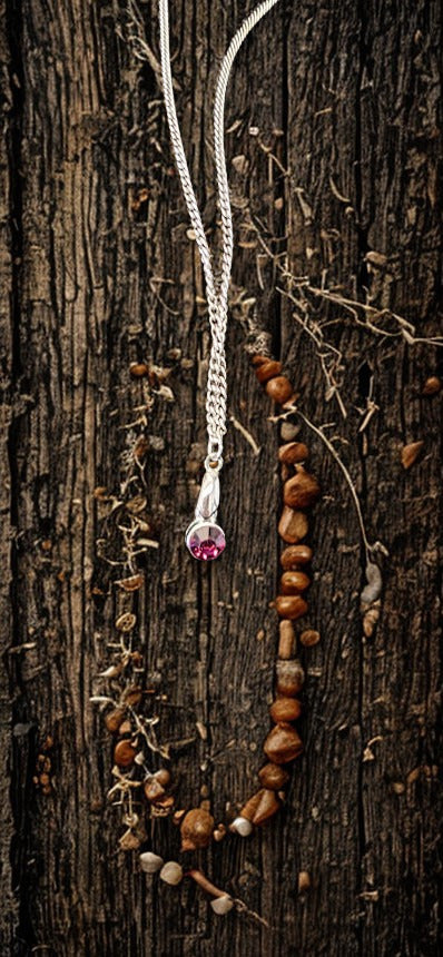 A rustic silver necklace with a small purple gemstone pendant, draped over a dark textured wood background alongside brown wooden beads and dried botanicals.