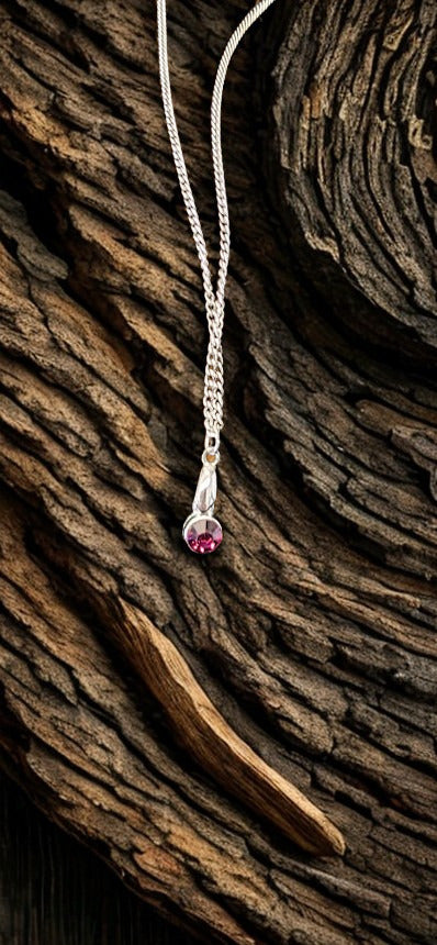 A silver-toned chain necklace with a teardrop pendant featuring a small PURPLE gemstone, displayed on a textured, dark brown wood background.