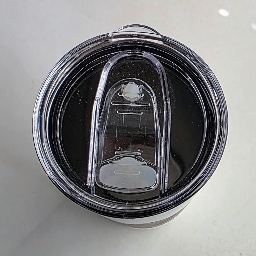 Top-down view of a clear plastic travel tumbler lid with a black rim and a transparent flip-up straw cover on a light gray surface.