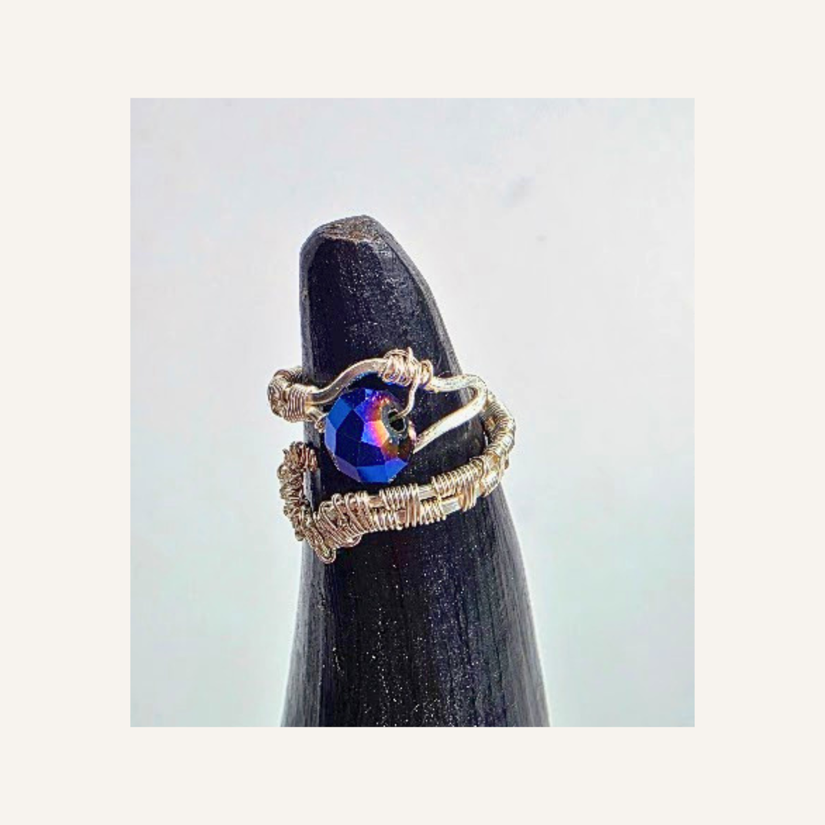 A handmade silver wire-wrapped ring with a central iridescent blue crystal, displayed on a black ring stand against a light gray background.