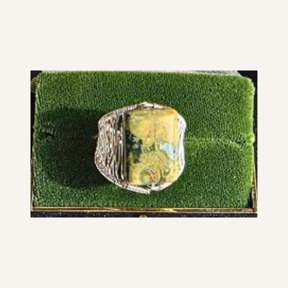 A silver-toned ring with a large, rectangular, marbled yellow-green stone, displayed on a green velvet ring box.