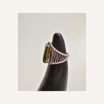 A silver-toned ring with a textured, openwork band and a polished olive-green stone, displayed on a dark wooden stand against a light gray background.