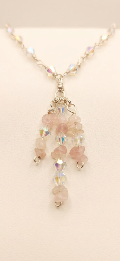 A silver-toned beaded necklace with a dangle pendant featuring light pink rose quartz beads and iridescent faceted beads, displayed on a neutral cream background.