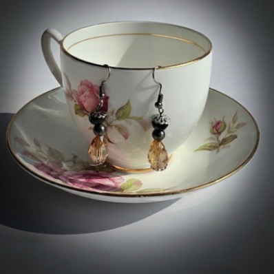 A pair of dangling earrings displayed on a white porcelain teacup and saucer with pink floral decoration and gold trim, set against a dark gray gradient background.
