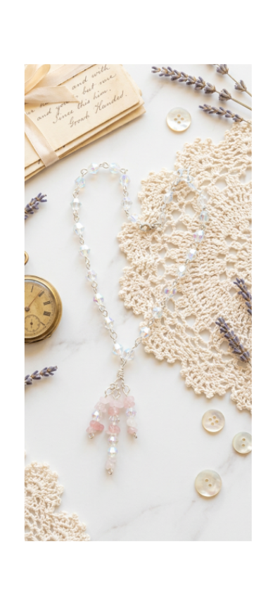A delicate beaded necklace with a pink tassel charm and silver-toned beads, styled in a flat lay on a white marble surface with lace, buttons, lavender, and a vintage pocket watch.