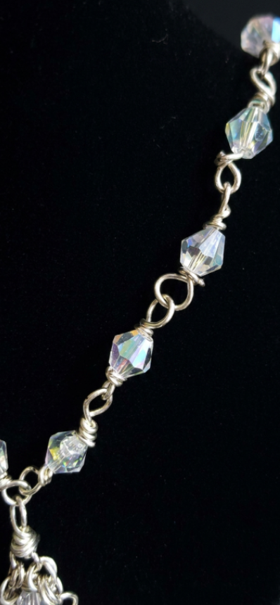 A close-up of a silver-toned beaded chain bracelet featuring faceted, iridescent clear beads with a metallic link design, set against a solid black background.