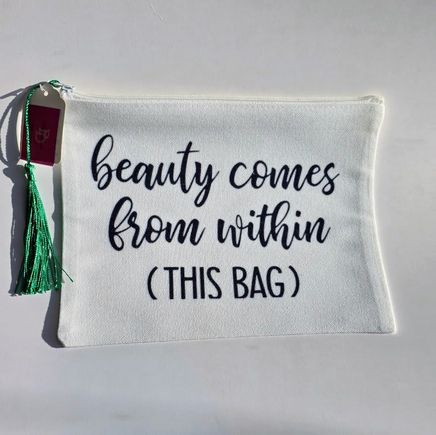 An off-white canvas cosmetic pouch with black embroidered text reading 'beauty comes from within (THIS BAG)'. It features a green tassel and a small maroon tag on the left side, set against a plain white background.