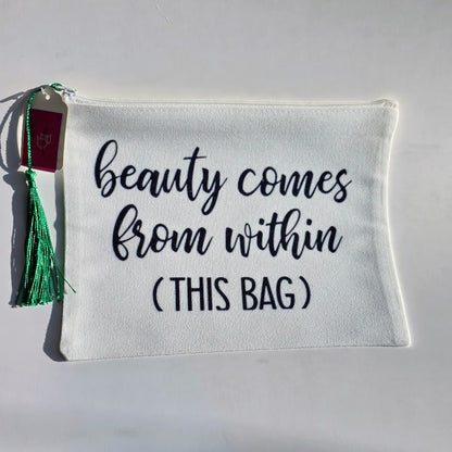 An off-white canvas cosmetic pouch with black embroidered text reading 'beauty comes from within (THIS BAG)'. It features a green tassel and a small maroon tag on the left side, set against a plain white background.