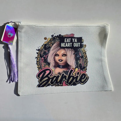 A white zippered pouch featuring a graphic print of a doll with pink hair, the text 'EAT YA HEART OUT' and 'Barbie', and a purple tassel with a holographic tag on a light gray background.