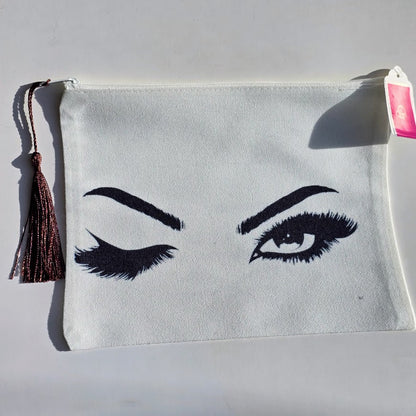 An off-white canvas pouch featuring a black embroidered design of one winking eye and one open eye with long lashes. The pouch has a brown tassel zipper pull and a pink price tag attached.